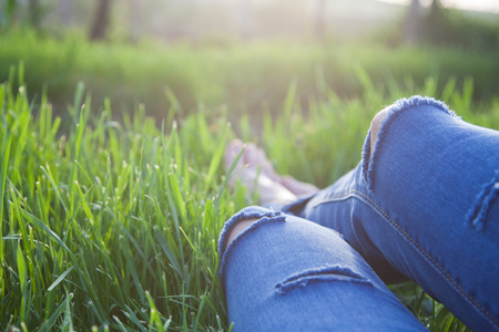 legs of hipster woman wearing torn jeans lying in green grass with smartphone and headsets enjoying sunset - summertime relaxationの写真素材