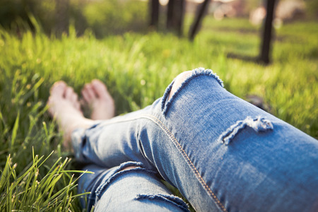 legs of hipster woman wearing torn jeans lying in green grass with smartphone and headsets enjoying sunset - summertime relaxationの写真素材