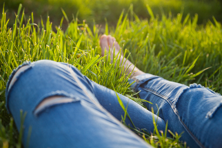 legs of hipster woman wearing torn jeans lying in green grass with smartphone and headsets enjoying sunset - summertime relaxationの写真素材
