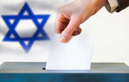 elections in Israel. Hand of a woman putting her vote in the ballot box. Waved Israel flag on background.の写真素材