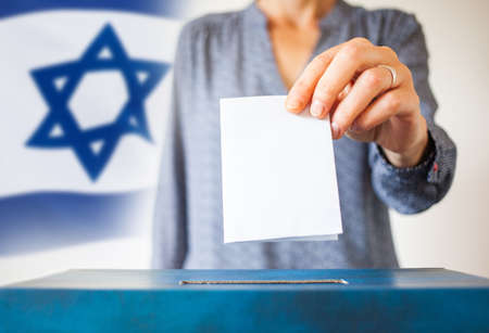 elections in Israel. Hand of a woman putting her vote in the ballot box. Waved Israel flag on background.の写真素材