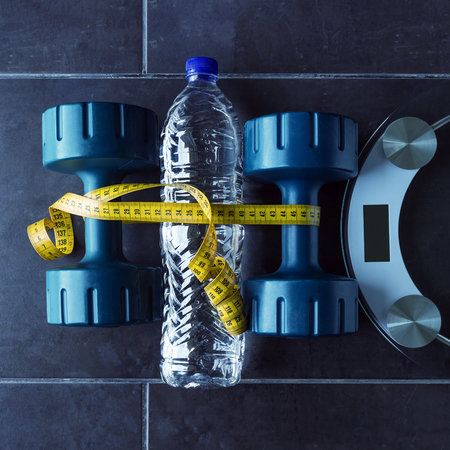 Dumbbells lie next to a bottle of pure mineral water and electronic scales. Yellow measuring tape unites them. Concept- lifestyle, sports and diet for weight loss.の写真素材