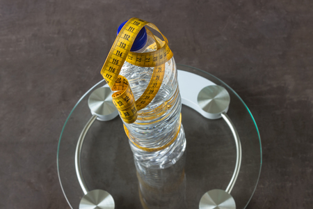 On the scales there is a bottle of pure mineral water with a yellow measuring tape. Selective focus. Concept- lifestyle, sports and diet for weight loss.の写真素材