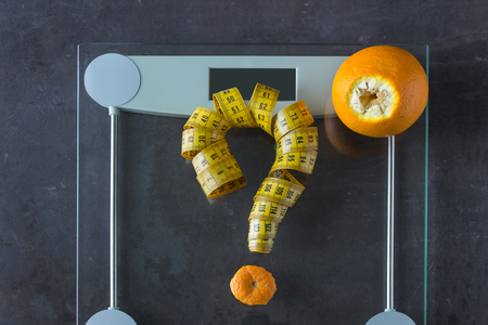 On the scales is measuring tape and fruit orange. Symbol-  question mark. Concept- lifestyle, sports and diet for weight loss.の写真素材