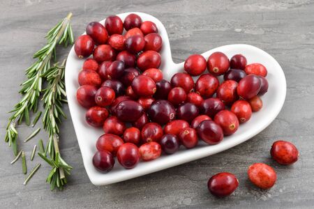 Heart healthy red Cranberries sit merrily on a heart shaped plate. Fresh organic rosemary graces the dark marble background to create a festive holiday or heart health concept.の写真素材