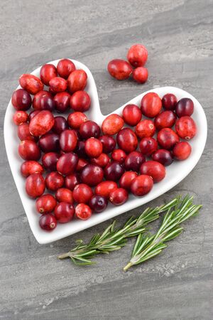 Heart healthy cranberries grace a heart shaped plate. A couple sprigs of fresh rosemary rest merrily next to the plate which sits on a dark marble background.の写真素材