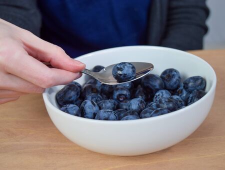 Closeup of caucasian woman dipping a spoon into a bowl of fresh organic blueberries. The bowl of blueberries rests on a rustic wooden table.の写真素材
