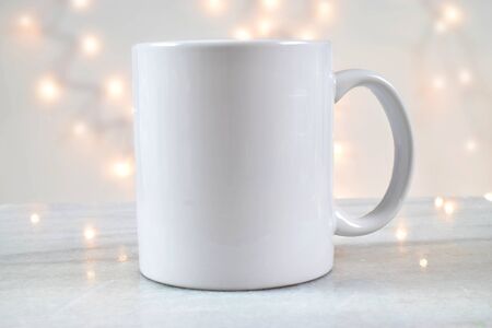 A blank 11 oz coffee cup rests delightfully on a marble background with glowing white Christmas lights in the background. Ready for you to place your own design on the coffee mug.の写真素材