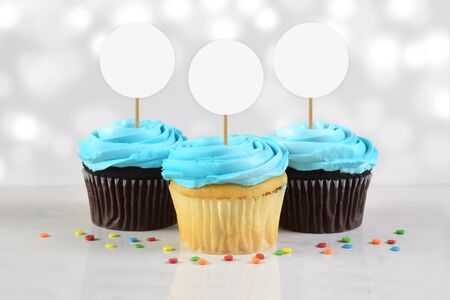 This cupcake topper mockup features three delicious blue frosted cupcakes atop a white marble background dusted with colorful sprinkles. The glowing white backround lights add a touch of class to this cupcake mock up.の写真素材