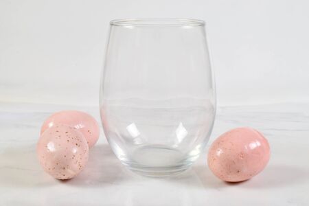 A luxurious no stem wineglass sits cheerfully on a white marble background. Three pretty pink speckled Easter eggs have the wine glass surrounded.の写真素材
