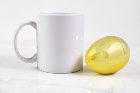 This Easter coffee cup mock up features an 11 ounce white coffee mug relaxing atop a white marble background. A gold Easter egg sits next to the mug adding a touch of class to this minimalist Easter mock-up.の写真素材