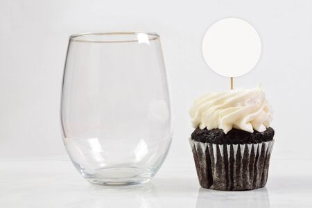 This cupcake topper and no stem wine glass mock up features an elegant stemless wineglass resting next to a tempting chocolate cupcake.の写真素材