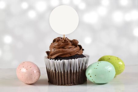 Gourmet chocolate cupcake rests on white marble background with colorful Easter eggs. 