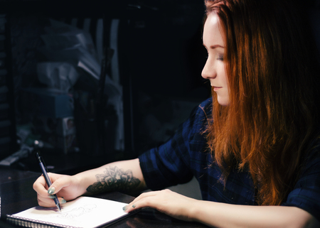 Artist designer jeweler girl draws a sketch of unusual jewelry ornaments with a pen on white paper. Home workshopの写真素材