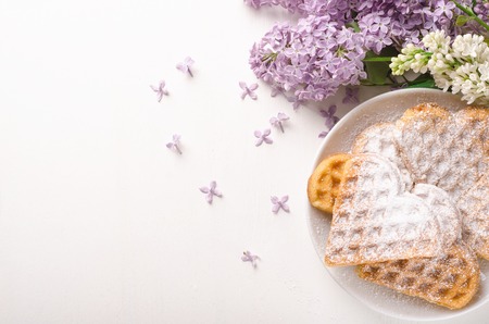 Homemade waffles heart sprinkled with powdered sugar on plate on a white table with flowers. Sweet pastry. Top viewの写真素材
