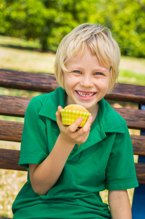 Cute happy school child eating a homemade cupcake outdoors on a park benchの写真素材