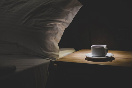 A cozy night scene with a white cup of coffee on a wooden bedside table next to a pillow and soft light, evoking comfort, warmth, and relaxation in a hotel or home bedroom.の写真素材
