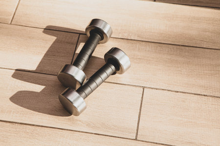 Pair of metal dumbbells on wooden floor in sunlight. Minimalist home fitness concept symbolizing self-training, healthy lifestyle, and daily workout motivation.の写真素材