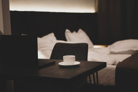 Cozy hotel room with a laptop and coffee cup on a desk, unmade bed in the background, creating a...
