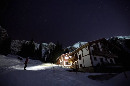 Man tourist in the mountains shine to a white chalet at night under a sky full of stars.