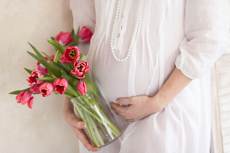 Hands of the pregnant woman on a stomach. Hands of the pregnant woman and spring flower. White dress. Mother's hands. Pink tulips.の写真素材