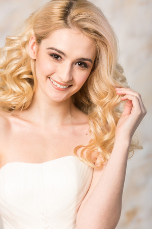 Portrait of the gorgeous smiling blonde bride with beautiful curly hair in a white wedding dress closeup on a light background.の写真素材