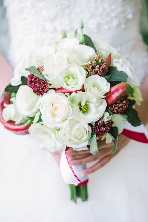 Bride in a white wedding dress holding a flowers bouquet from white rose and red fresh peppers in hands. Wedding bridal bouquet on a white background close up.の写真素材