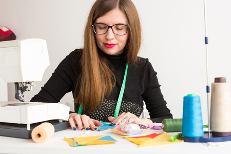 needlework in the workshop of a young woman, a tailor - pretty tailor in glasses at work with pieces of colored cloth on the table with threads, fabrics, needles and sewing machine. Hobby concept.の写真素材