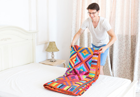 handsome smiling young man folds double a color patchwork quilt on big bed in the white interior....