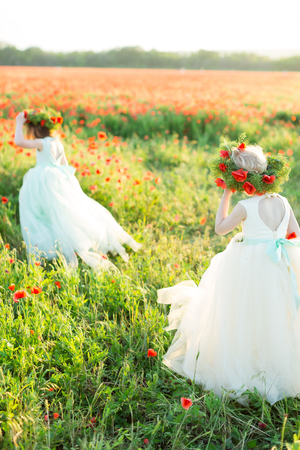 little girl model, poppies, summer fashion concept - on the field of poppies two young fairies for a walk in white elegant dresses, both girls are wearing garlands of red flowers, they go awayの写真素材