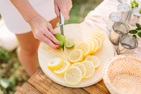 picnic, people, food, summer, nature concept - young slender girl in a white dress with a knife cut lemon into round slices, lie on a plate of lemons and green lime, dishes, glasses on wooden tableの写真素材