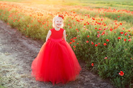 kids fashion, happy childhood, spring, nature concept - laughing beautiful blonde girl with flower on head in bright red dress standing on the rural road near the field of poppiesの写真素材