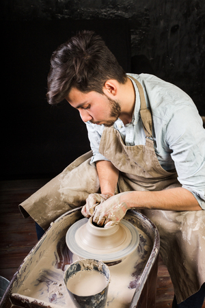 potter, workshop, ceramics art concept - closeup on working potters wheel with raw clay and mans hands, young brunette male sculpt a utensil with fingers, master in apron, Mexican, Hispanic, Latinoの写真素材