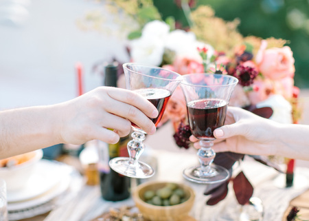 Close -up Of Couple Hands Toasting Red Wine on festively table with flowers and fresh food background. birthday, event, wedding concept. Man and woman drinking red wine.の写真素材