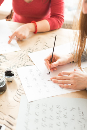 inspiration, patience, lettering concept. two women wearing grey and red sweatshirts creating various fonts with help of special equipment on the desk covered brown paper for sketchingの写真素材