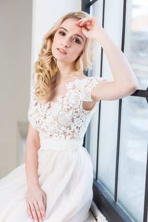 fashionable gown, beautiful blonde model, bride hairstyle and makeup concept - pretty young woman in white wedding dress sitting on the windowsill indoors, romantic girl posing in the studioの写真素材