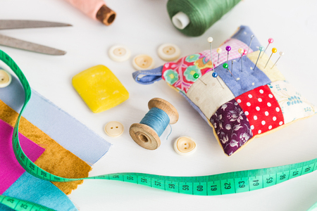 closeup sewing tools , patchwork, tailoring and fashion concept - working environment on a white table, thread spools, buttons, meter, pincushion, scissors, pieces of colored patchwork fabric, soap