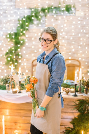 decorating, planting, celebrating concept. there is young lovely woman, worker of the restaurant, she is wearing comfortable denim shirt and holding transparent vase with flowersの写真素材