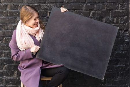 fashion, femininity, advertisment concept. in front of black brick wall there is adorable loughing woman that is holding blackboard for drawing with colorful chalksの写真素材