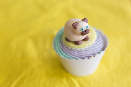 sweets, treatment, confection concept. on the yellow background of tablecloth there is marvelous cake in paper cup decorated with figure of cat that is lying on the top of whipped creamの写真素材