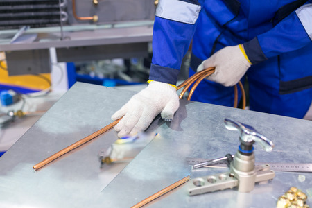 manufacturing, technology, factory concept. there is man who dressed in special blue uniform and gloves for protecting when he is working with welding, he is holding hank of copper thin tubeの写真素材