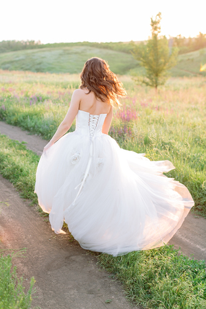 wedding, marriage, freedom concept. in the light of the setting sun wonderful bride in white puffy dress is running through the field with small wildflowers and a tree in the distanceの写真素材