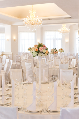 design, party, holidays concept. in the light hall of restaurant decorated for the feast there are tables and chairs that are covered with white textile and everywhere amount of flowersの写真素材
