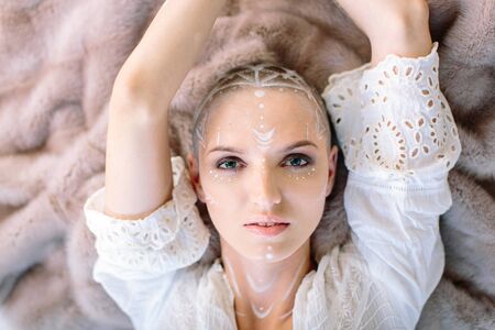 From above mysterious woman with white paint art on face and neck raising hands up and looking at cameraの写真素材