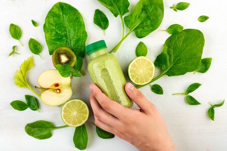 A man's hand grab a bottle of light green smoothie detox lying on a white background surrounded by kiwi, apple, lime, spinach. The concept of propper male nutrition Flat lay, top viewの写真素材
