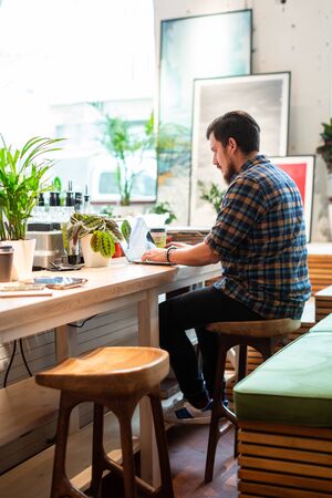 Man working on laptop in coffee shop. Casual modern man sitting at wooden counter in coffee shop and...