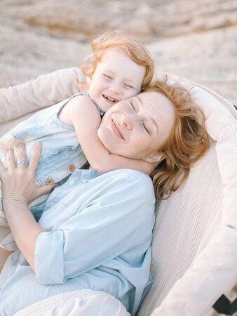adorable lovely child and her mother with red hairs dressed in light blue shirt, they are hugging each other and laughingの写真素材