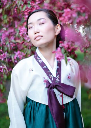 Concept of asian beauty. Portrait of marvelous korean woman with closed eyes and smile, asian girl wearing silk hanbok and standing nearby with sakura tree treeの写真素材