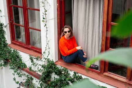 A woman wearing sunglasses and casual clothes sits on the open window with her back leaned on frame...