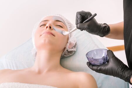 Process of applying a silver cosmetic mask with brush to female client head skin, on a white background. Concept of facial cosmetic treatments at the spa.の写真素材
